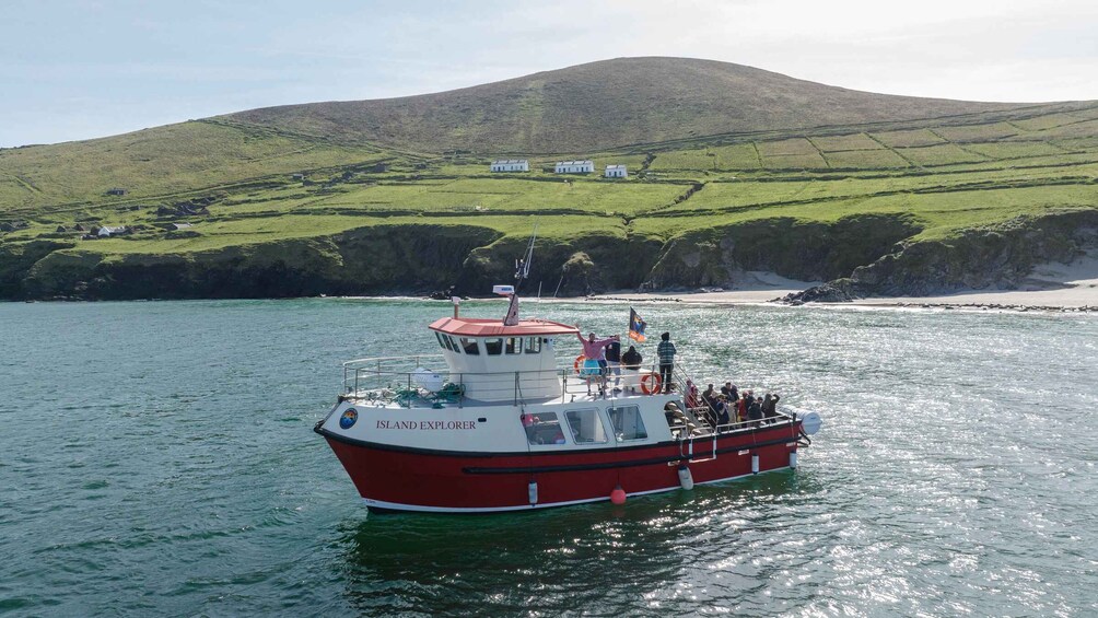 A red and white boat named 'Island Explorer' on calm waters with a green hill and houses in the background. 2