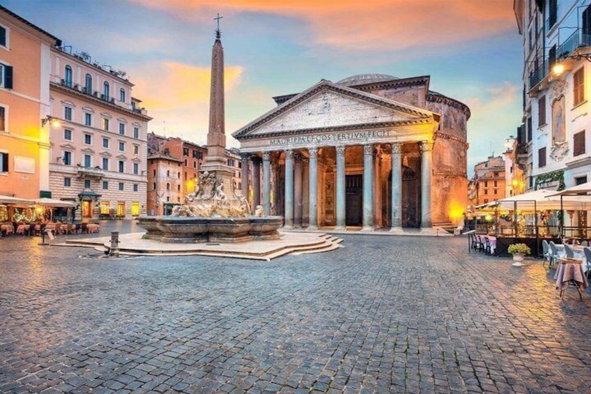 A cobblestone square with a classical fountain, a tall obelisk, and a neoclassical building with columns. 1