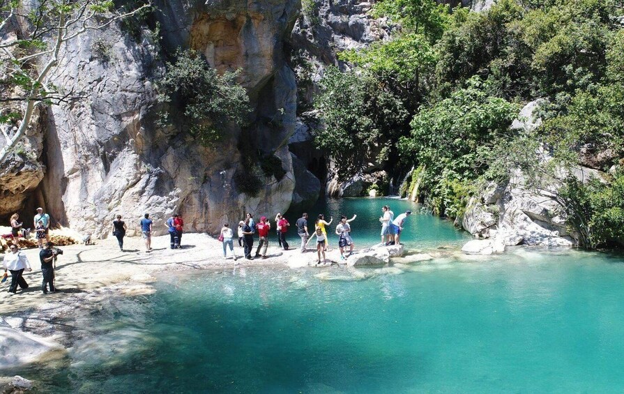 A group of people standing on rocks by a clear turquoise pool surrounded by rocky cliffs and greenery. 4