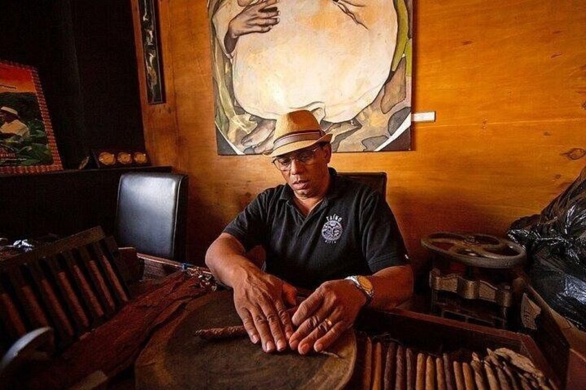 A person seated at a wooden table with a cigar in hand, a hat on their head, and a watch on their wrist. 3