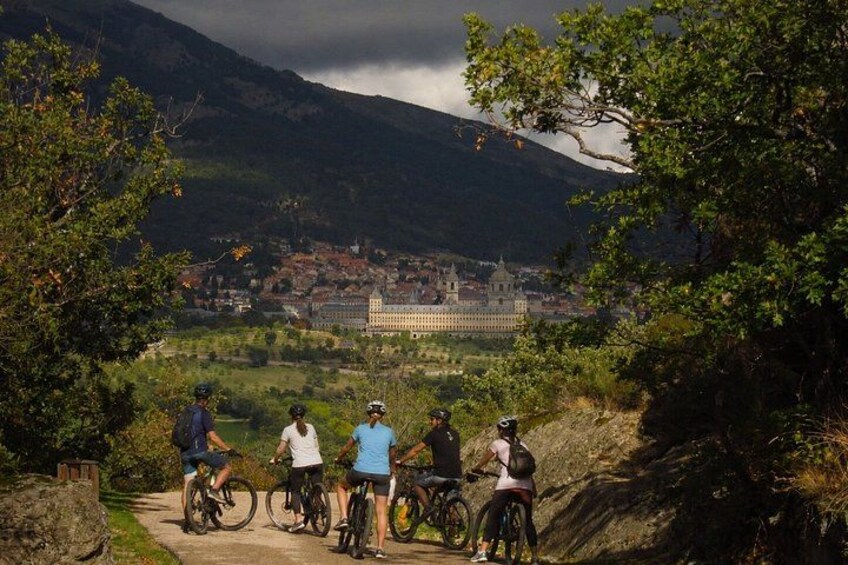A group of cyclists on a path with a mountainous landscape and a large building in the background. 1