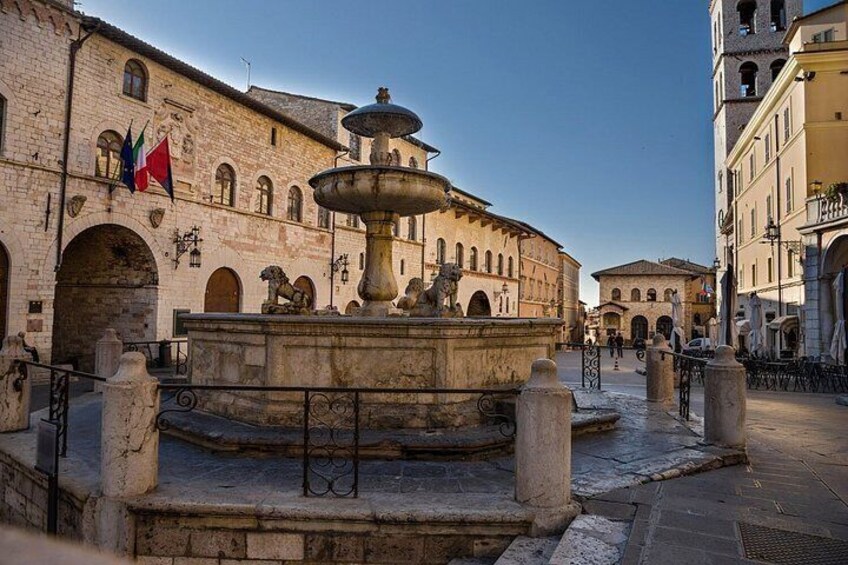 A historic square with a central fountain, stone buildings, and flags. 4