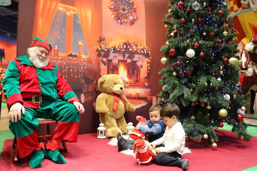 A person in a red and green outfit sitting next to a Christmas tree and a fireplace. 3