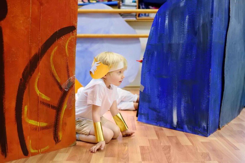 A child sitting on the floor, wearing a crown and gold arm bands, in a room with wooden flooring and colorful walls. 2
