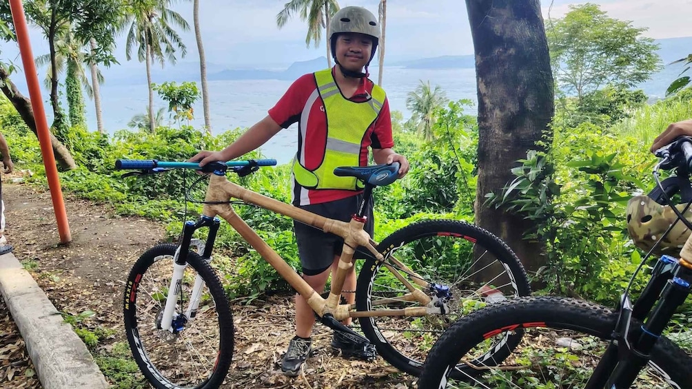 A person standing next to a mountain bike with a helmet, in a natural setting with trees and a view of the sea in the background. 2