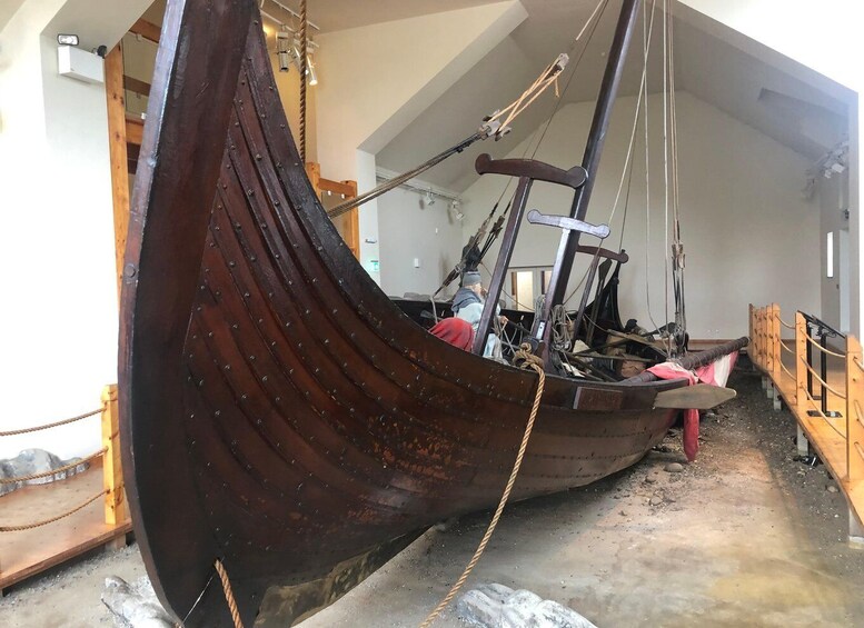 A large wooden ship with a curved bow, ropes, and a metal mast inside a building. 5