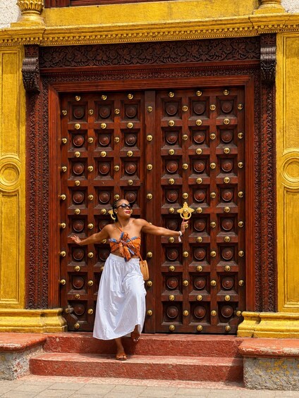 A person standing in front of a large ornate wooden door with multiple locks, wearing a white skirt and a colorful top. 1