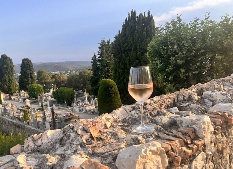 A glass of white wine on a stone ledge overlooking a cemetery with tombstones and trees. 3