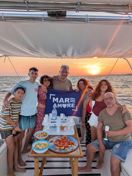 A group on a boat with a sunset in the background, a banner reading 'MARAMORE,' and a table with food and drinks. 1