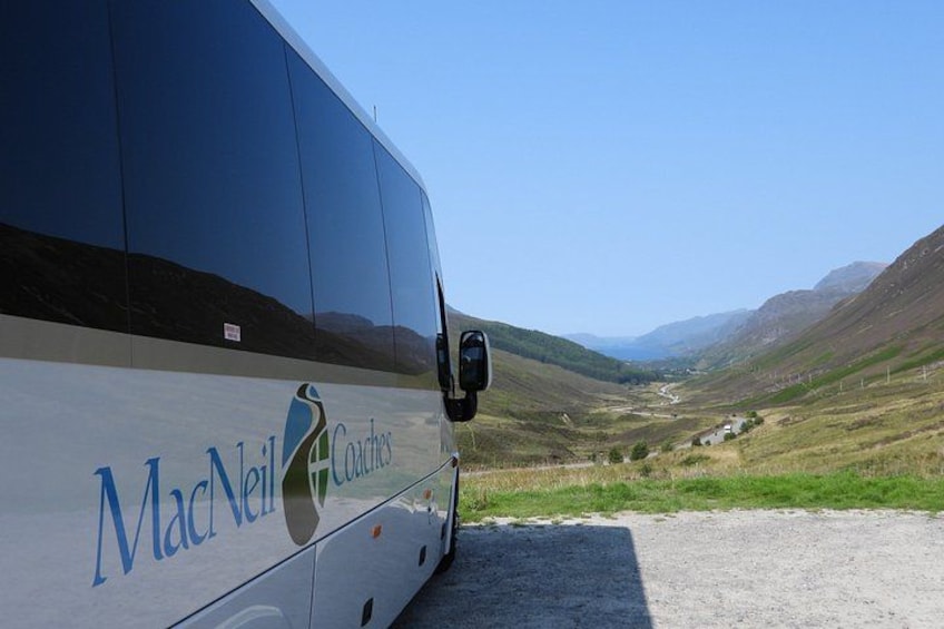 A bus with 'MacNeil Coaches' written on the side, parked on a gravel road with a scenic view of a valley and hills in the background. 2