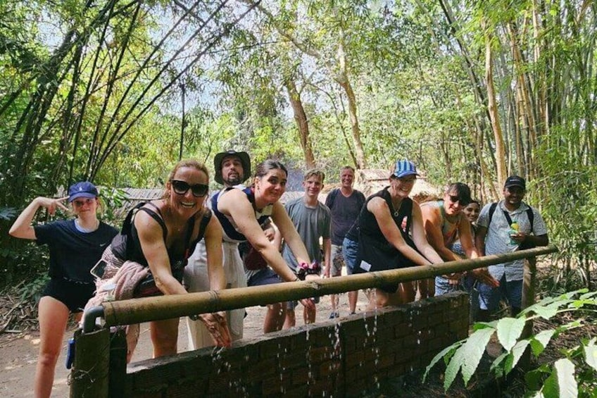 A group of people in a forested area, some standing on a wooden bridge, others on a dirt path. 2
