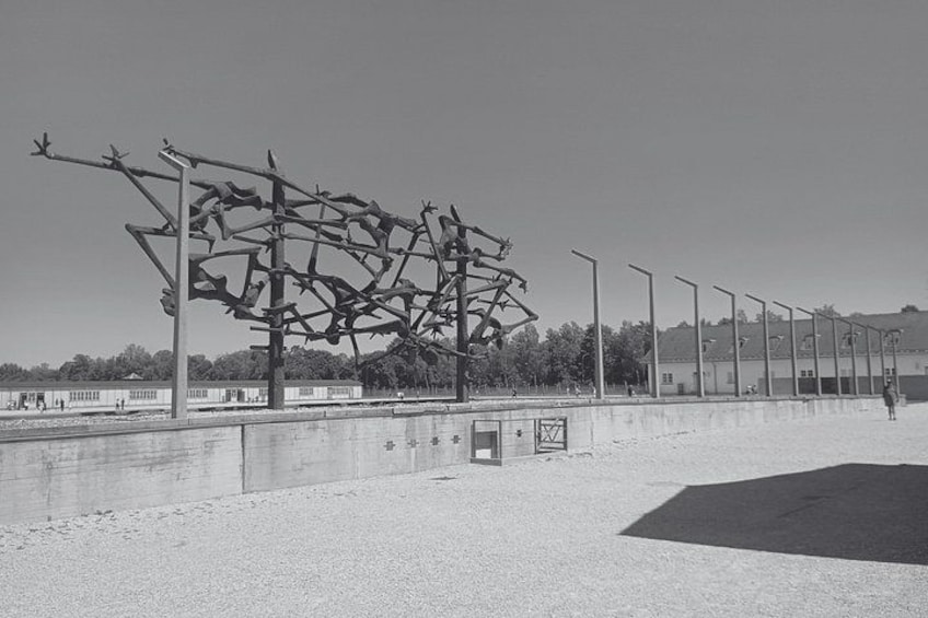 A large metal sculpture on a concrete platform by a waterfront, with a row of street lamps and a person walking in the distance. 3