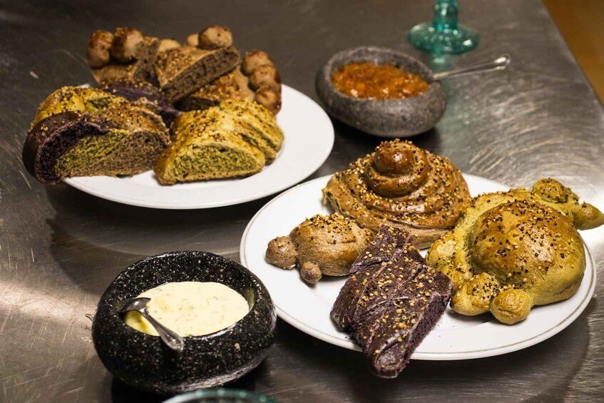 Variedad de productos horneados en platos, incluyendo pan y pastelería, con un bol de mantequilla y un recipiente de especias al fondo. 1