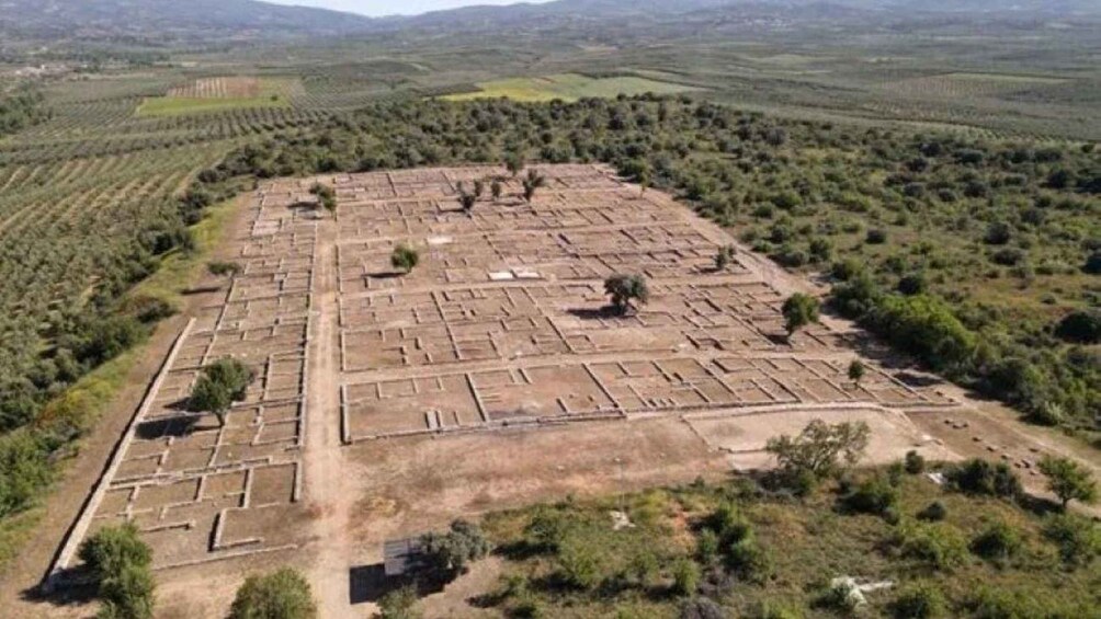 An aerial view of a large, rectangular archaeological site with grid-like patterns of ruins, surrounded by a rural landscape with scattered trees and fields. 3