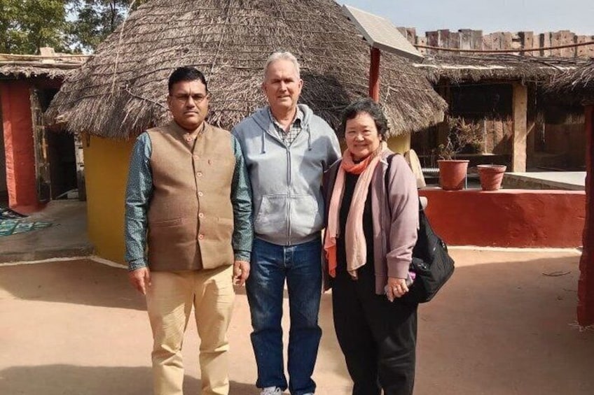 Three people standing in front of a traditional thatched-roof hut with a red wall and potted plants. 3