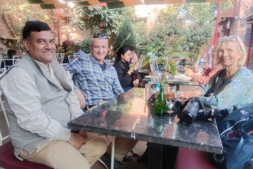 People sitting at an outdoor cafe with tables, chairs, and greenery in the background. 4