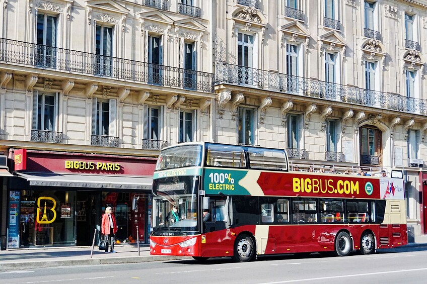 Punainen, kaksikerroksinen bussi, jonka kyljessä on tekstit ”100 % SÄHKÖINEN” ja ”BIGBUS.COM”, on pysäköitynä koristeellisten parvekkeiden ja ”BIGBUS PARIS” -kyltin koristaman liikkeen eteen. 3