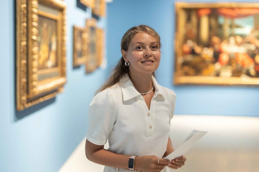 En person i en hvid skjorte holder papirer i et kunstgalleri med indrammede malerier på væggene. 5