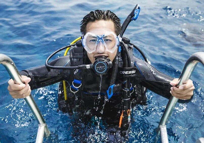 A person in a black wetsuit with a scuba diving tank and regulator, holding onto metal handrails in the water. 1