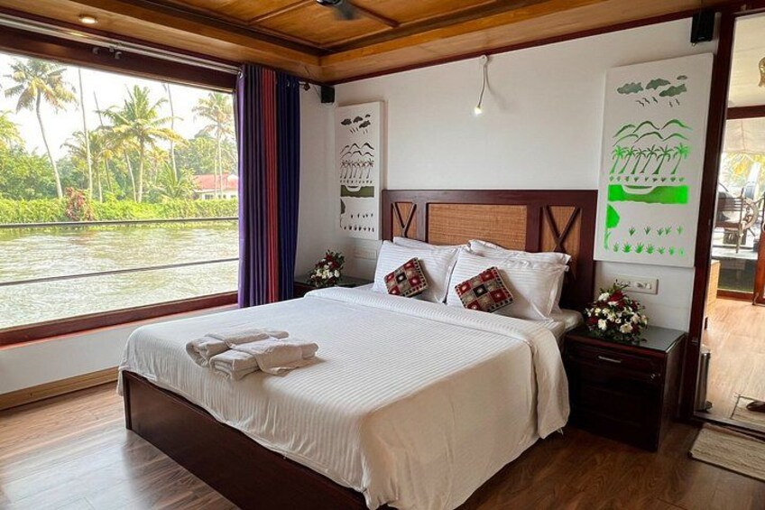 A large bed with white linens, a wooden headboard, and decorative pillows. There is a nightstand with a lamp and a flower arrangement. A window with a view of palm trees and water is visible. 5