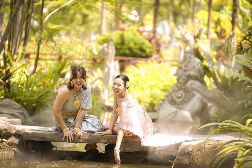 Two people in traditional attire sitting on a wooden bridge over a body of water, surrounded by lush greenery and a stone statue. 4