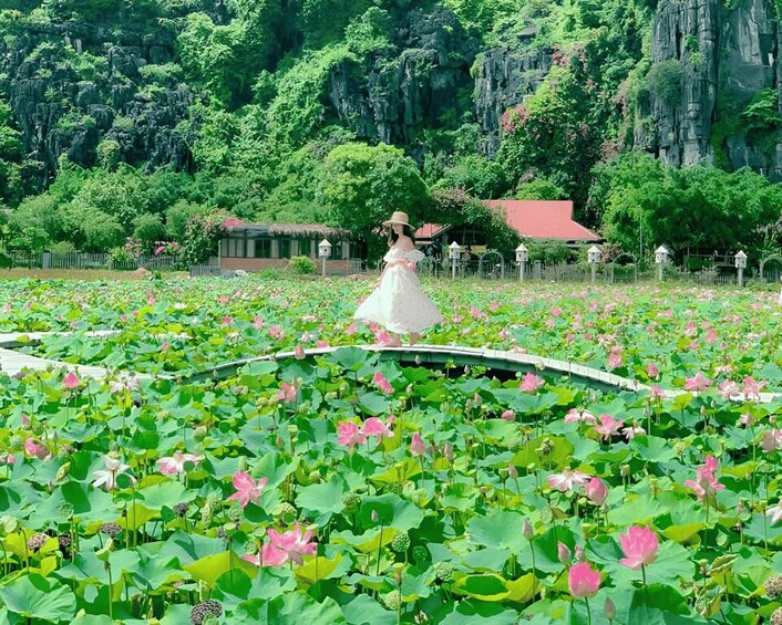 A woman in a white dress walking on a lotus pond surrounded by lush greenery and a red-roofed building. 5