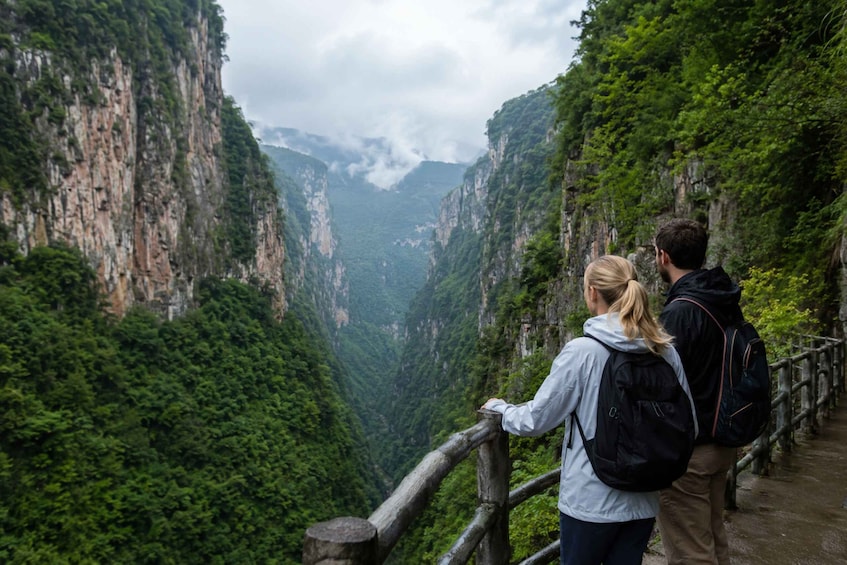 Two people standing on a wooden walkway, overlooking a deep valley with steep cliffs and dense greenery. 1
