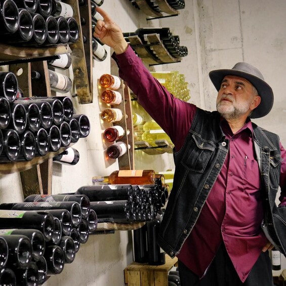 A person in a hat and vest pointing at a wine rack with various bottles. 4