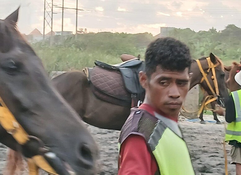 A person standing next to a horse with a saddle, another horse in the background, and a person in a reflective vest. 2