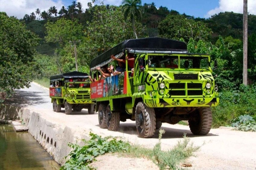 A row of green off-road vehicles parked on a concrete path beside a body of water, surrounded by lush greenery. 4