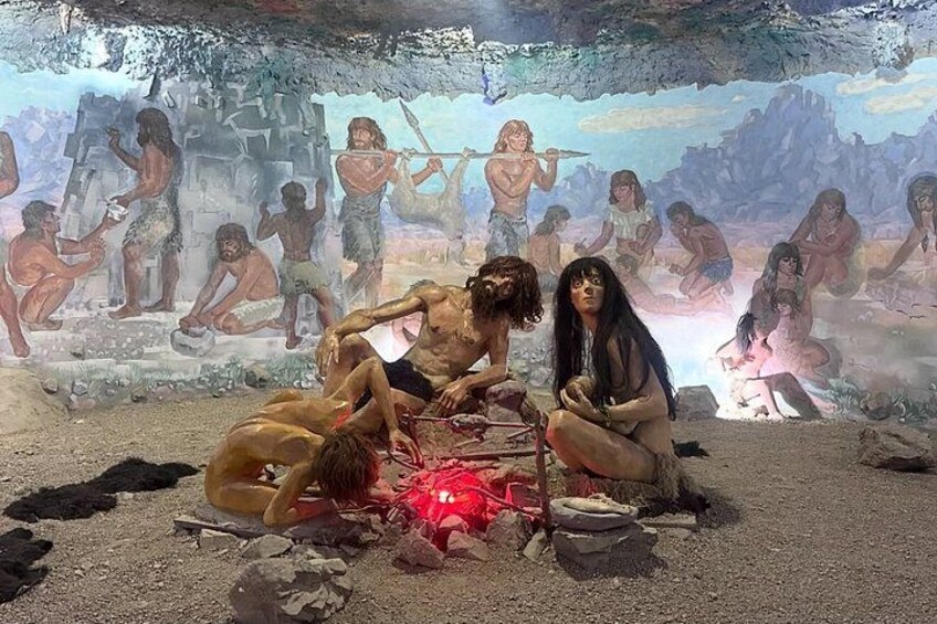A prehistoric scene with figures around a fire, cave paintings in the background, and a rocky terrain. 2