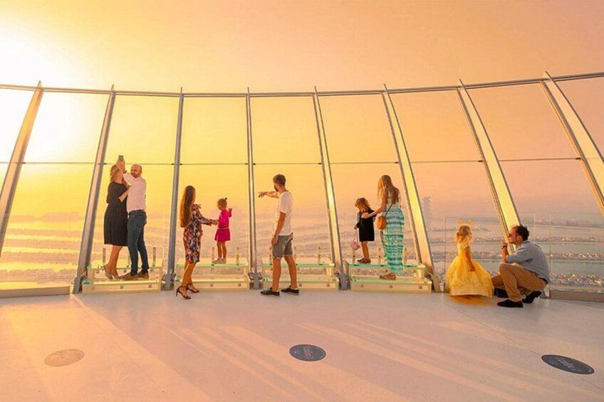 People standing and sitting on a glass floor, overlooking a cityscape during sunset. 2