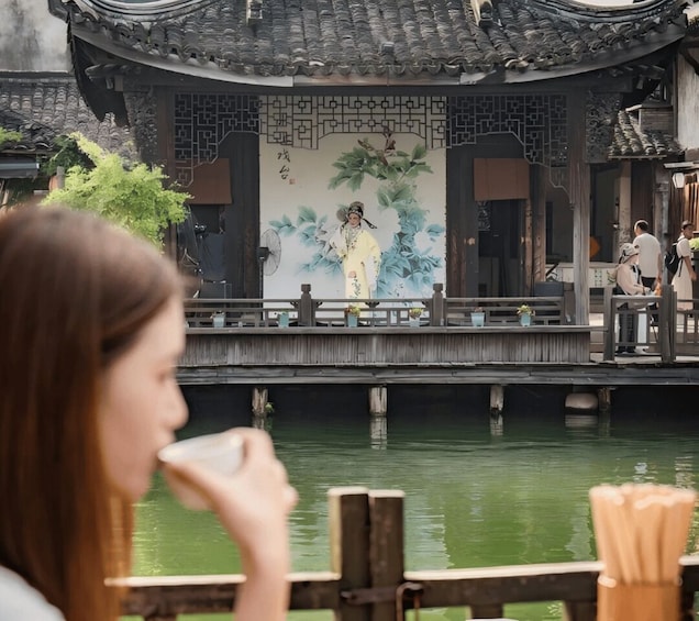 A traditional Chinese building with a mural, a wooden bridge over water, and a person sitting at a table. 1