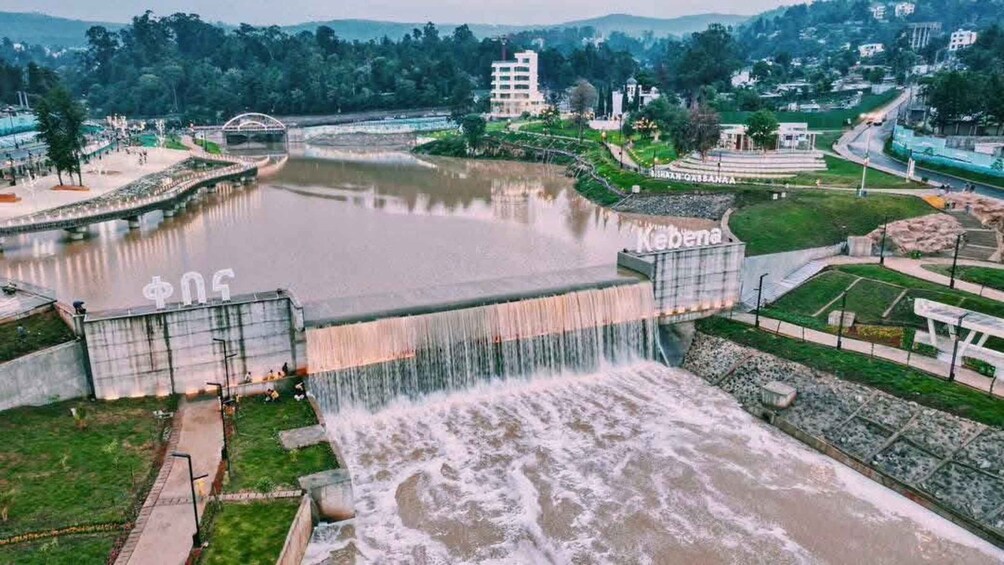 A dam with water flowing over it, a bridge, and buildings in the background. 4
