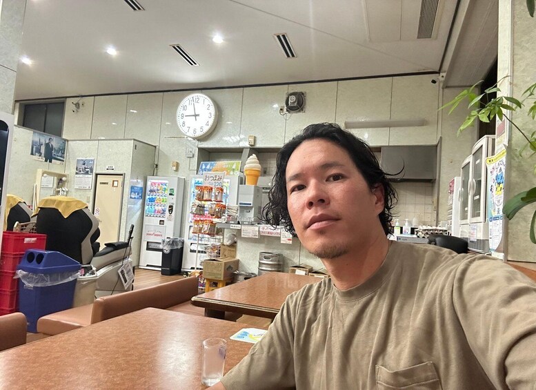 A person seated at a table in a restaurant with a clock on the wall, vending machines, and a plant in the background. 5