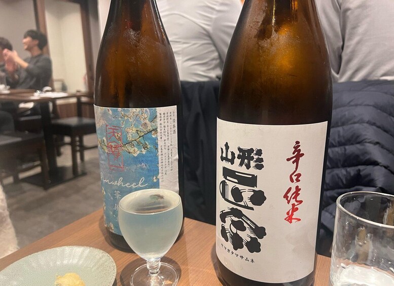 Two bottles of Japanese sake with labels, a glass of sake, and a plate with food on a wooden table in a restaurant. 4