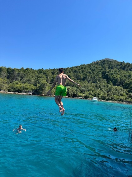 A person jumping into turquoise water with a boat and forested hill in the background. 1