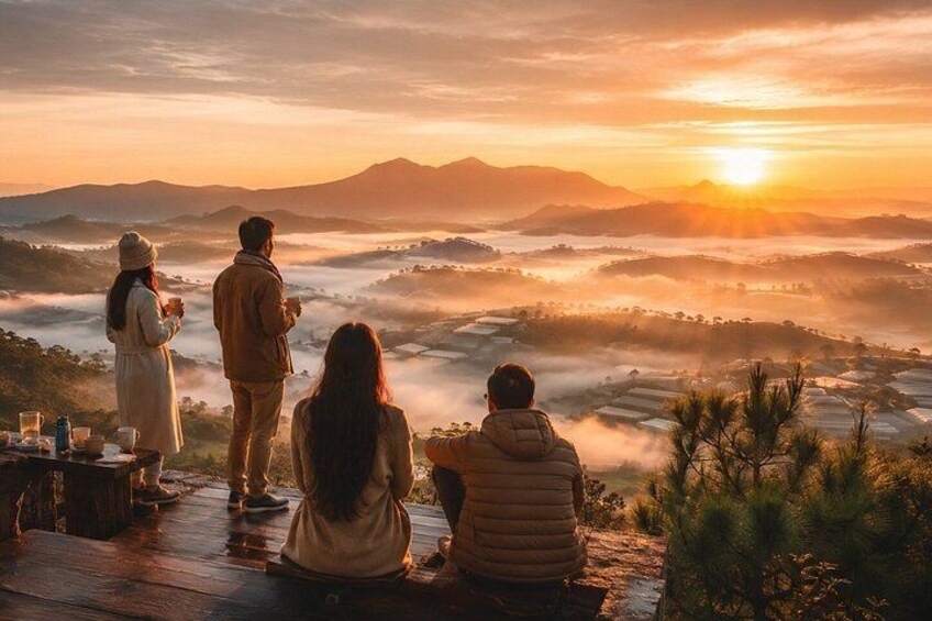 A group of people sitting and standing on a wooden deck overlooking a valley with mist-covered hills and a setting sun. 1