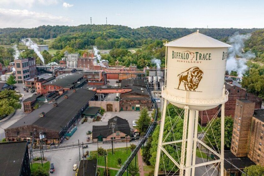 A water tower with 'Buffalo Trace Distillery' written on it, surrounded by industrial buildings and a green landscape in the background. 1