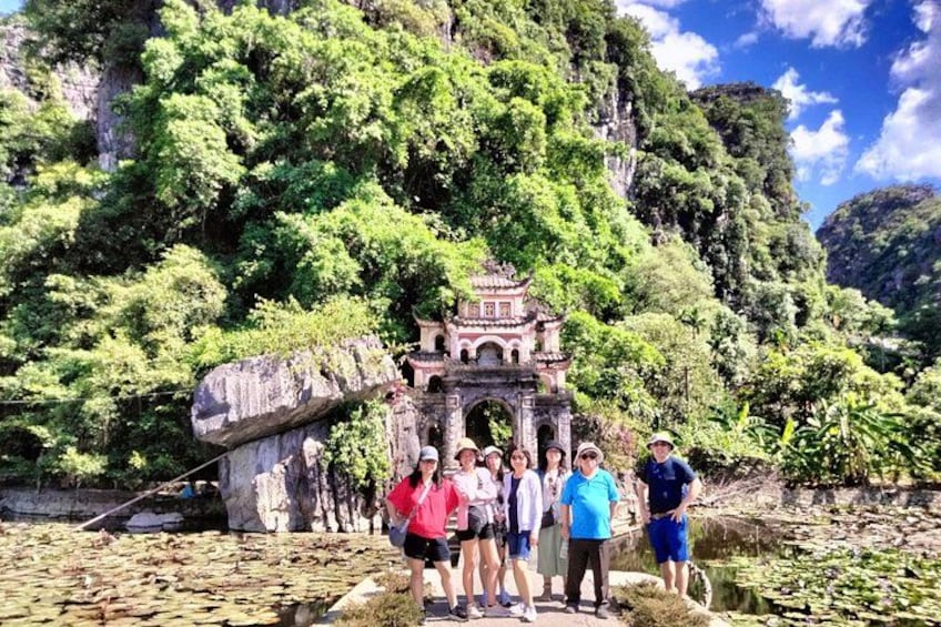 A group of people standing in front of a stone structure with a traditional roof, surrounded by lush greenery and a rocky cliff. 3