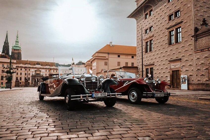 A vintage car with a trailer parked on a cobblestone street, historic buildings, and a clock tower in the background. 4