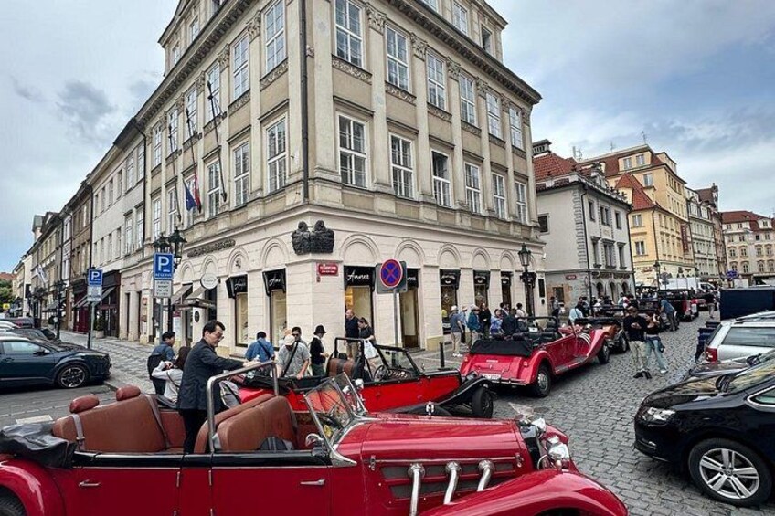 Vintage cars parked on a cobblestone street, a building with flags, and pedestrians. 2