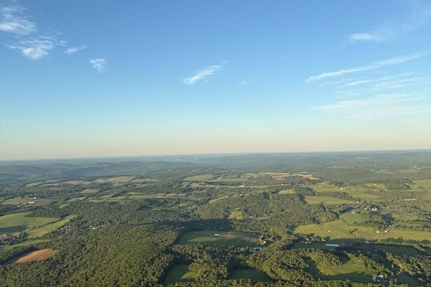 A landscape with rolling hills, patches of forest, and scattered fields under a clear sky. 2
