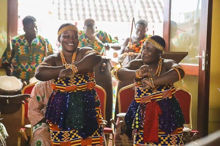 Two individuals in colorful traditional attire, seated indoors with musical instruments in the background. 1