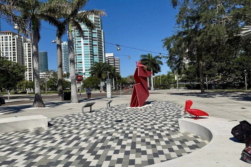 A plaza with a checkered floor, palm trees, a red sculpture, and modern buildings in the background. 1