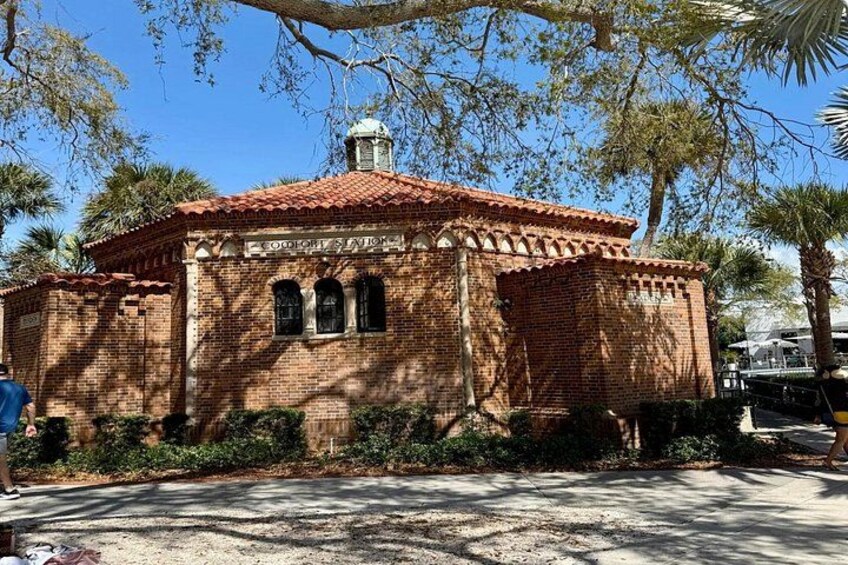 A brick building with a red-tiled roof, surrounded by greenery and palm trees. 4