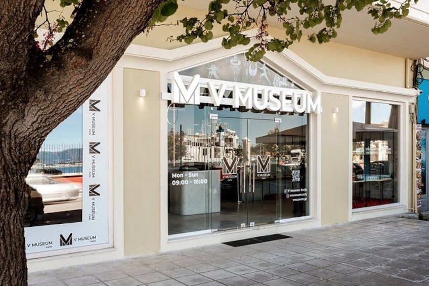 A museum with a large glass facade, signage, and a tree in the foreground. 1
