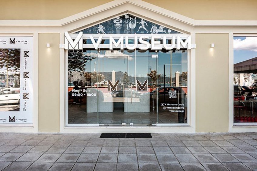 The MV Museum entrance with a large glass facade, a tiled pavement, and a clear sky. 3