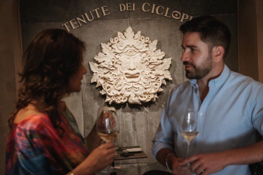 Two people holding wine glasses in front of a wall with a decorative carving and the text 'TENUTE DEI CICLOPI'. 4