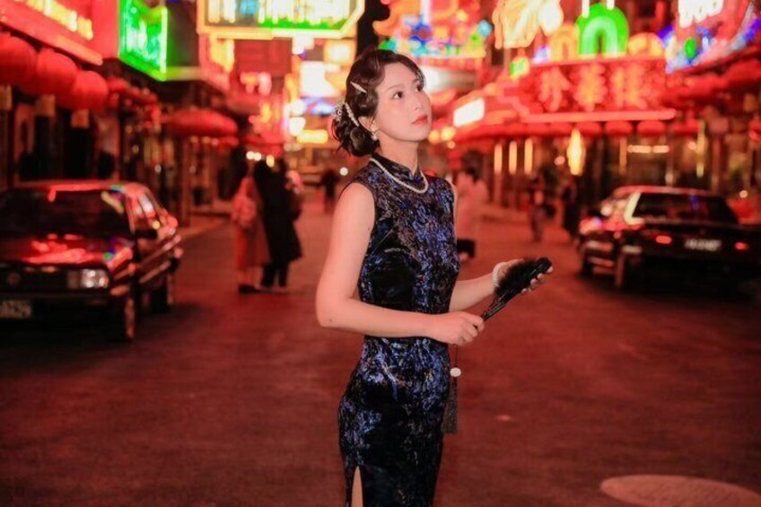 A person in a patterned dress holding a microphone on a brightly lit street with neon signs. 2